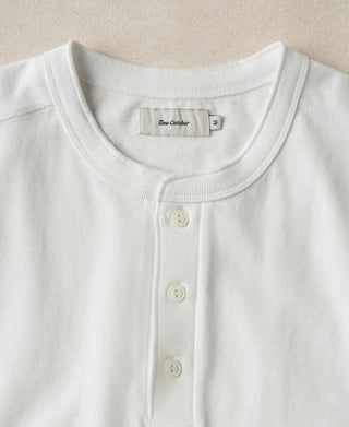 A close-up of a white Henley shirt by Blue Clover, featuring a ribbed crew neck and three-button placket, laid flat against a neutral beige background.