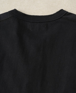 Close-up of the back of a black crewneck sweatshirt, showing the ribbed collar and a small rectangular patch on the inside neck label, set against a light beige textured background.