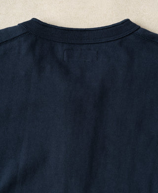Close-up of the back of a dark navy knit sweater, showing the ribbed crew neck and a small rectangular patch on the upper back, laid flat on a light beige textured surface.