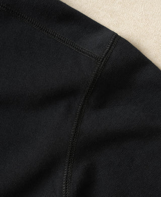 Close-up detail of a black knit garment showing a shoulder seam and ribbed fabric texture against a light neutral background.